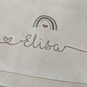May include: A cream-colored knit blanket featuring an embroidered design of a small rainbow with a heart underneath. Below the rainbow, the name Elisa is stitched in a brown cursive script, which begins with a small heart and ends with a long line.