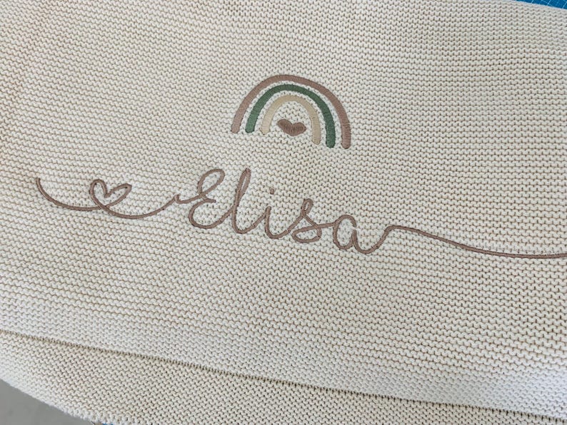 May include: A cream-colored knit blanket featuring an embroidered design of a small rainbow with a heart underneath. Below the rainbow, the name Elisa is stitched in a brown cursive script, which begins with a small heart and ends with a long line.