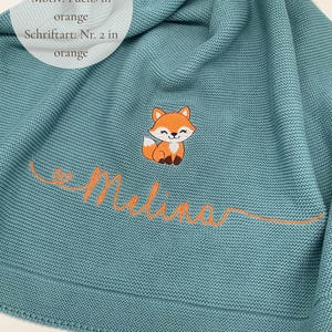 May include: A smoke blue knitted blanket featuring an embroidered orange fox and the name Melina in orange cursive script. The text on the blanket reads: Deckenfarbe: Rauchblau, Motiv: Fuchs in orange, Schriftart: Nr. 2 in orange.