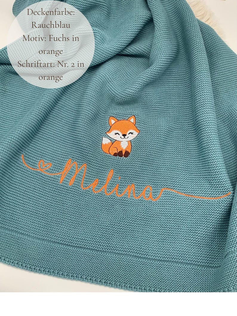 May include: A smoke blue knitted blanket featuring an embroidered orange fox and the name Melina in orange cursive script. The text on the blanket reads: Deckenfarbe: Rauchblau, Motiv: Fuchs in orange, Schriftart: Nr. 2 in orange.