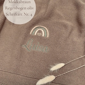 May include: A mocha brown knitted blanket featuring an embroidered design of a rainbow in olive and beige tones with a small heart at the center. Below the rainbow, the name Luisa is embroidered in olive green script. Text reads: Mokkabraun, Regenbogen oliv, Schriftart Nr. 4.