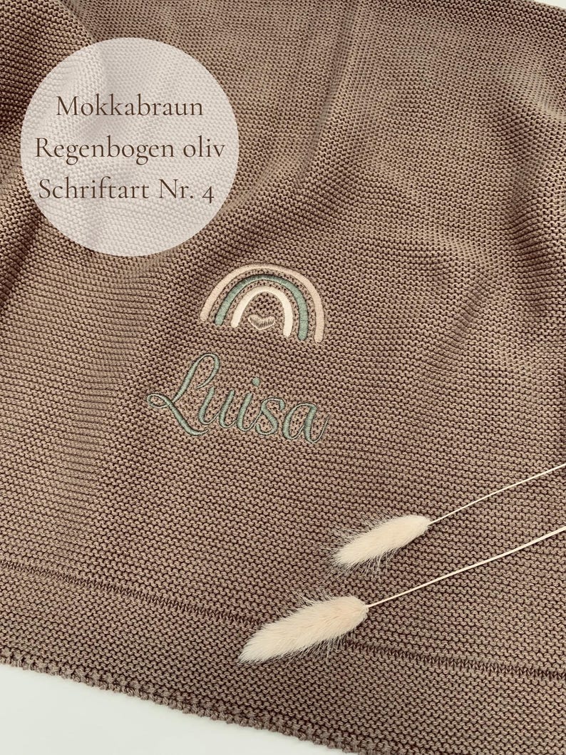 May include: A mocha brown knitted blanket featuring an embroidered design of a rainbow in olive and beige tones with a small heart at the center. Below the rainbow, the name Luisa is embroidered in olive green script. Text reads: Mokkabraun, Regenbogen oliv, Schriftart Nr. 4.