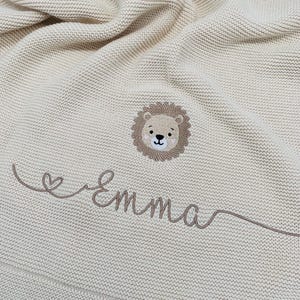 May include: A cream-colored knitted blanket featuring a central embroidered lion face with a brown mane. Below the lion, the name Emma is stitched in brown cursive script, accompanied by a small heart icon integrated into the lettering design.
