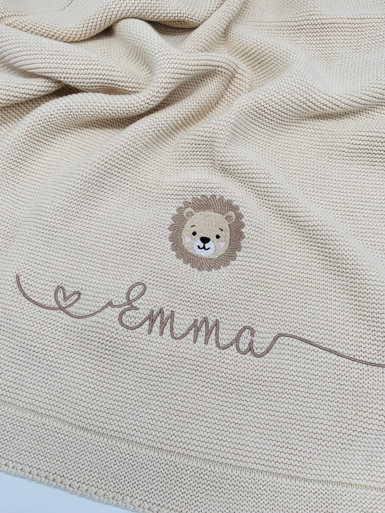 May include: A cream-colored knitted blanket featuring a central embroidered lion face with a brown mane. Below the lion, the name Emma is stitched in brown cursive script, accompanied by a small heart icon integrated into the lettering design.