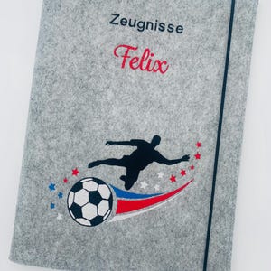 May include: A light gray felt document folder featuring the embroidered text Zeugnisse and Felix in red and black. The design includes a black silhouette of a soccer player, a soccer ball, and small red, white, and blue stars with a curved swoosh.