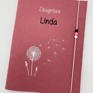 Könnte beinhalten: Ein staubrosa Filzordner mit den Wörtern "Zeugnisse Linda" in Weiß und Schwarz. Auf der Vorderseite ist ein weißes Pusteblumen-Design aufgedruckt. Ein rosa Gummiband mit schwarzen, rosa und weißen Perlen sichert den Ordner.