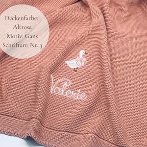 May include: A dusty pink knitted blanket featuring an embroidered white goose with a pink bow around its neck. The name Valerie is embroidered in white cursive script below the goose. Text on the image reads Deckenfarbe: Altrosa, Motiv: Gans, Schriftart: Nr. 3.