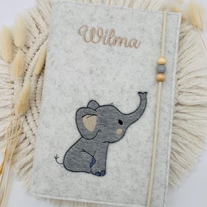 May include: A light gray felt notebook cover featuring an embroidered gray elephant appliqu&eacute; and the name Wilma stitched in brown thread. A vertical elastic band with two wooden beads and one gray bead secures the cover.