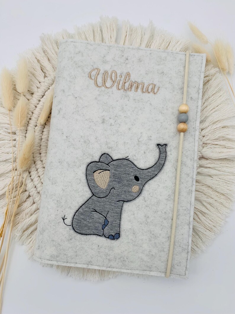 May include: A light gray felt notebook cover featuring an embroidered gray elephant appliqu&eacute; and the name Wilma stitched in brown thread. A vertical elastic band with two wooden beads and one gray bead secures the cover.