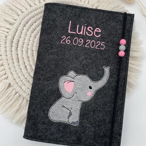May include: A dark gray felt notebook cover featuring the embroidered name Luise and the date 26.09.2025 in pink thread. A gray elephant applique with pink ear and cheek details is centered below. A black elastic band with three beads secures the cover.