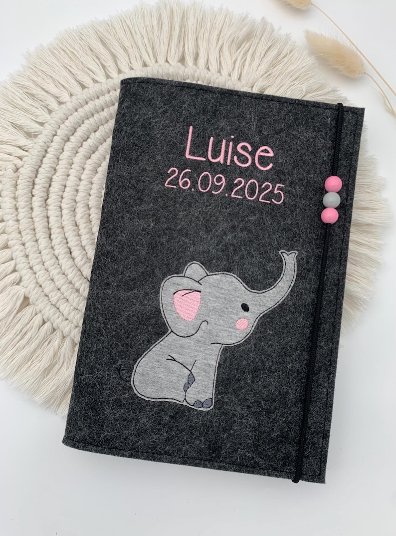 May include: A dark gray felt notebook cover featuring the embroidered name Luise and the date 26.09.2025 in pink thread. A gray elephant applique with pink ear and cheek details is centered below. A black elastic band with three beads secures the cover.