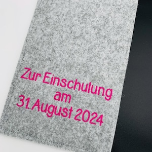 Könnte beinhalten: Grauer Filzordner mit schwarzem Filzklappe. Der Ordner ist mit pinkfarbener Stickerei versehen, die "Zur Einschulung am 31. August 2024" lautet.