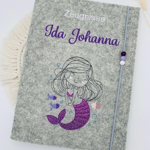 Op de afbeelding: Een grijze vilten map met de geborduurde tekst Zeugnisse en de naam Ida Johanna in het paars. Onder de tekst staat een geborduurde zeemeermin met een paarse staart, omringd door kleine vissen en bubbels. Een grijs elastiek met drie kralen.