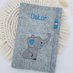 May include: A gray felt notebook cover featuring an embroidered elephant with blue accents and the name Oskar in blue thread. A vertical gray elastic band with two blue beads and one gray bead secures the cover, which has stitched edges.