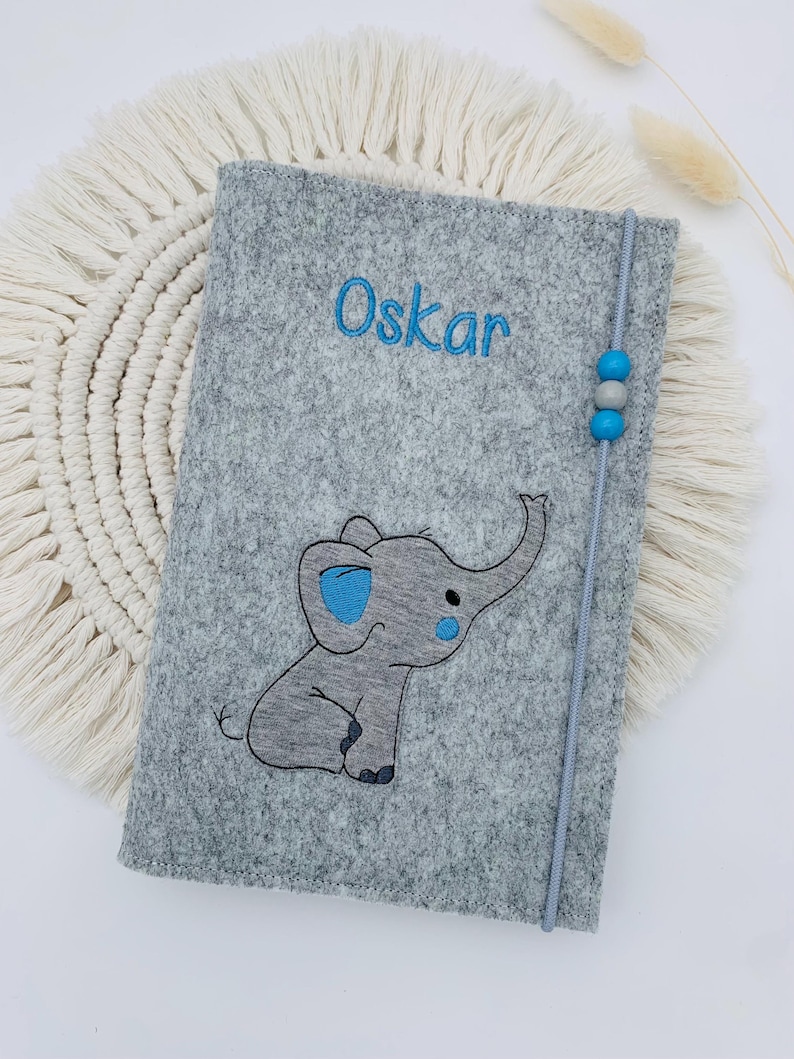 May include: A gray felt notebook cover featuring an embroidered elephant with blue accents and the name Oskar in blue thread. A vertical gray elastic band with two blue beads and one gray bead secures the cover, which has stitched edges.