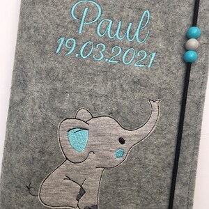 May include: A gray felt notebook cover featuring the embroidered name Paul and the date 19.03.2021 in light blue thread. An embroidered gray elephant with light blue accents sits below the text. A black elastic cord with two blue and one gray bead secures the cover.
