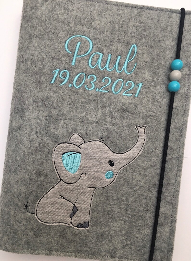 May include: A gray felt notebook cover featuring the embroidered name Paul and the date 19.03.2021 in light blue thread. An embroidered gray elephant with light blue accents sits below the text. A black elastic cord with two blue and one gray bead secures the cover.