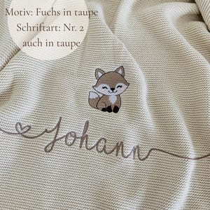 May include: A beige knitted blanket featuring an embroidered taupe fox graphic and the name Johann in cursive script. Text on the image reads: Decke: Natur, Motiv: Fuchs in taupe, Schriftart: Nr. 2, auch in taupe. The blanket has a textured knit.