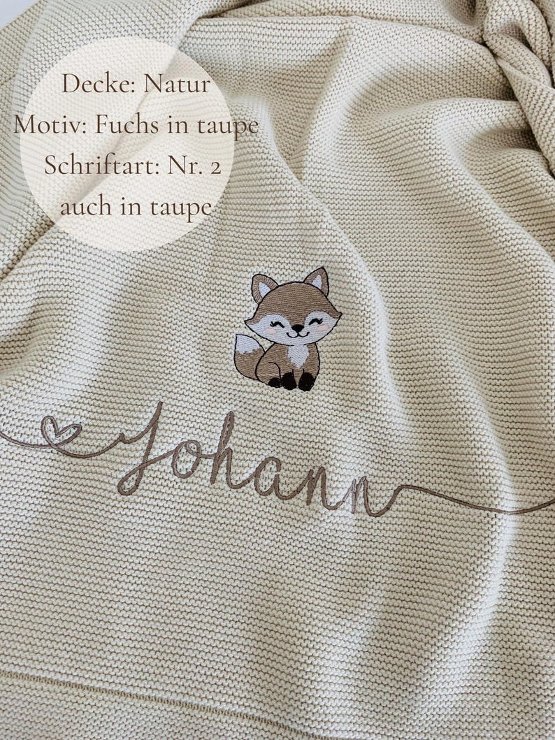May include: A beige knitted blanket featuring an embroidered taupe fox graphic and the name Johann in cursive script. Text on the image reads: Decke: Natur, Motiv: Fuchs in taupe, Schriftart: Nr. 2, auch in taupe. The blanket has a textured knit.