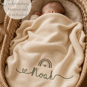 May include: A cream-colored knitted baby blanket featuring an embroidered design of a rainbow and a heart above the name Noah in green cursive script. The blanket is draped over a sleeping infant inside a woven wicker bassinet.
