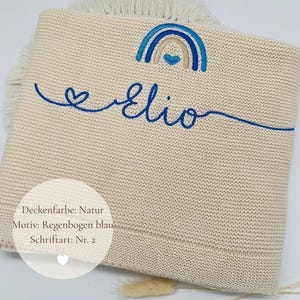 May include: A beige knitted blanket featuring a blue embroidered rainbow with a heart and the name Elio in cursive script. The blanket has a fringe edge and includes a circular overlay with the text Deckenfarbe: Natur, Motiv: Regenbogen blau, Schriftart: Nr. 2.