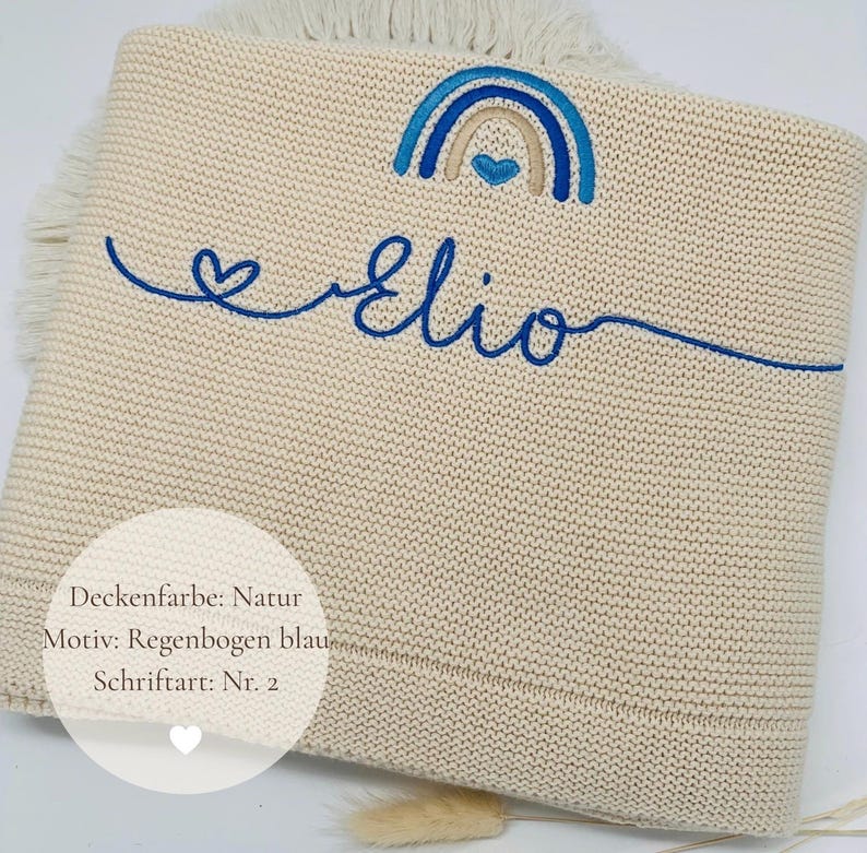 May include: A beige knitted blanket featuring a blue embroidered rainbow with a heart and the name Elio in cursive script. The blanket has a fringe edge and includes a circular overlay with the text Deckenfarbe: Natur, Motiv: Regenbogen blau, Schriftart: Nr. 2.