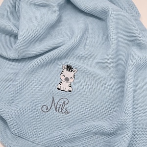 May include: A light blue knitted blanket featuring an embroidered graphic of a small zebra with black stripes and pink cheeks. Below the zebra, the name Nils is embroidered in a dark gray cursive script font. The fabric has a textured knit pattern.