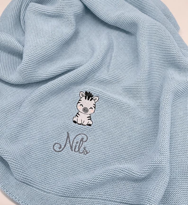 May include: A light blue knitted blanket featuring an embroidered graphic of a small zebra with black stripes and pink cheeks. Below the zebra, the name Nils is embroidered in a dark gray cursive script font. The fabric has a textured knit pattern.