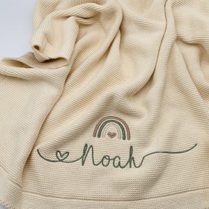 May include: A cream-colored knit blanket featuring a textured garter stitch pattern. Embroidered on the fabric is a small rainbow in muted earth tones with a heart in the center, followed by the name Noah in cursive green thread.