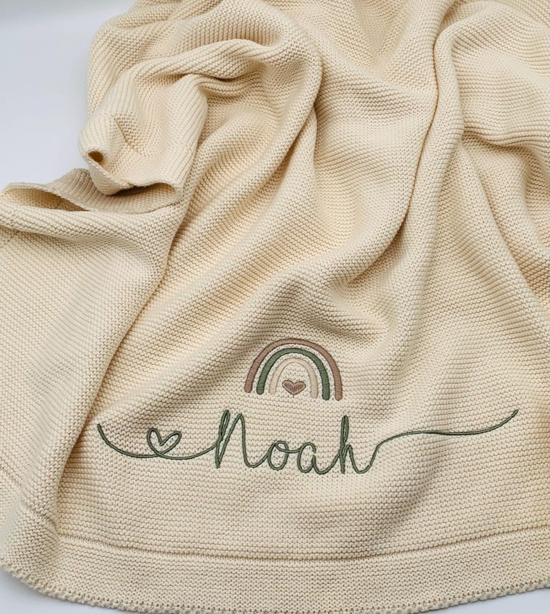 May include: A cream-colored knit blanket featuring a textured garter stitch pattern. Embroidered on the fabric is a small rainbow in muted earth tones with a heart in the center, followed by the name Noah in cursive green thread.