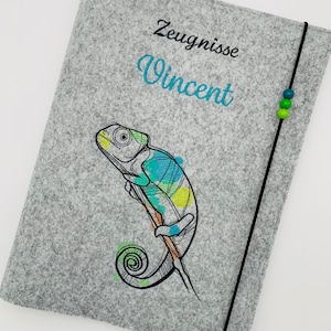Könnte beinhalten: Grauer Filzordner mit schwarzem Bandverschluss. Der Ordner ist mit einem gestickten Chamäleon mit grünen, blauen und gelben Akzenten verziert. Der Text "Zeugnisse Vincent" ist auf den Ordner gestickt.
