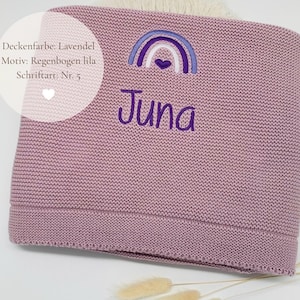 May include: A lavender knitted blanket featuring an embroidered purple rainbow with a small heart at its center and the name Juna stitched in purple thread below the rainbow. The text on the image reads Deckenfarbe: Lavendel, Motiv: Regenbogen lila, Schriftart: Nr. 5.