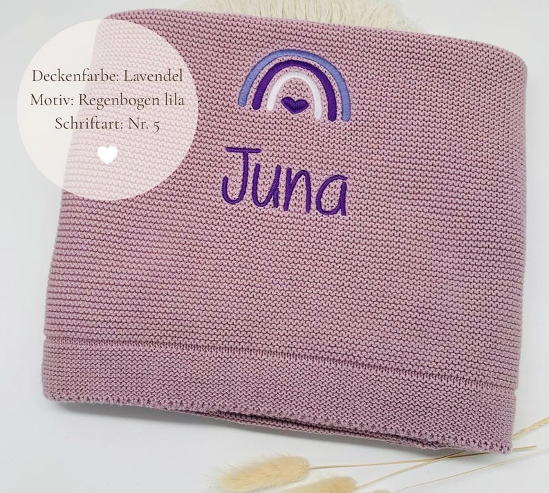 May include: A lavender knitted blanket featuring an embroidered purple rainbow with a small heart at its center and the name Juna stitched in purple thread below the rainbow. The text on the image reads Deckenfarbe: Lavendel, Motiv: Regenbogen lila, Schriftart: Nr. 5.