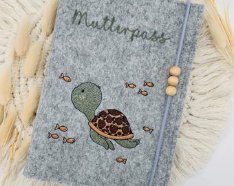 May include: A grey felt cover for a Mutterpass featuring an embroidered green turtle with a brown patterned shell, surrounded by small orange fish. The cover includes a light blue elastic closure band adorned with three round wooden beads.