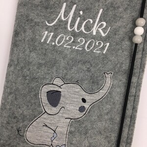 May include: A gray felt notebook cover featuring an embroidered elephant graphic, the name Mick, and the date 11.02.2021 in white thread. A black elastic band with three small gray and white beads secures the side of the cover.