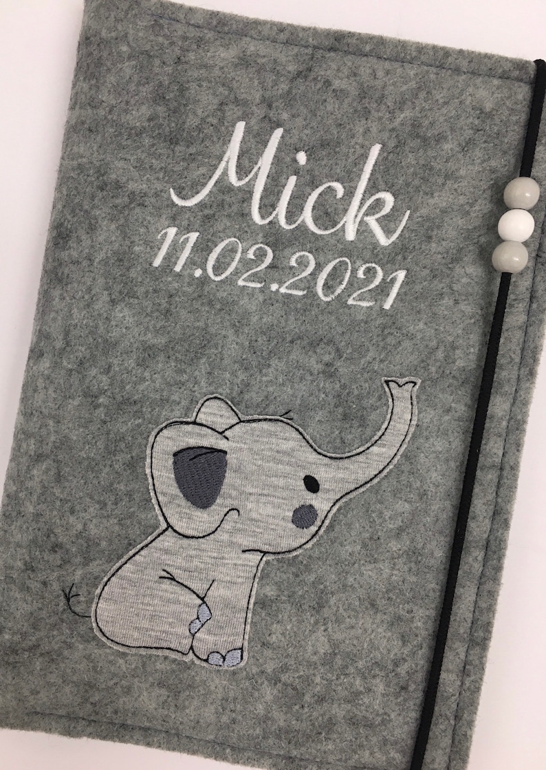 May include: A gray felt notebook cover featuring an embroidered elephant graphic, the name Mick, and the date 11.02.2021 in white thread. A black elastic band with three small gray and white beads secures the side of the cover.