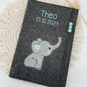 May include: A dark gray felt notebook cover featuring an embroidered light gray elephant with light blue accents. The cover is personalized with the name Theo and the date 13.10.2025 in light blue thread. A gray elastic band with three small beads secures the cover.