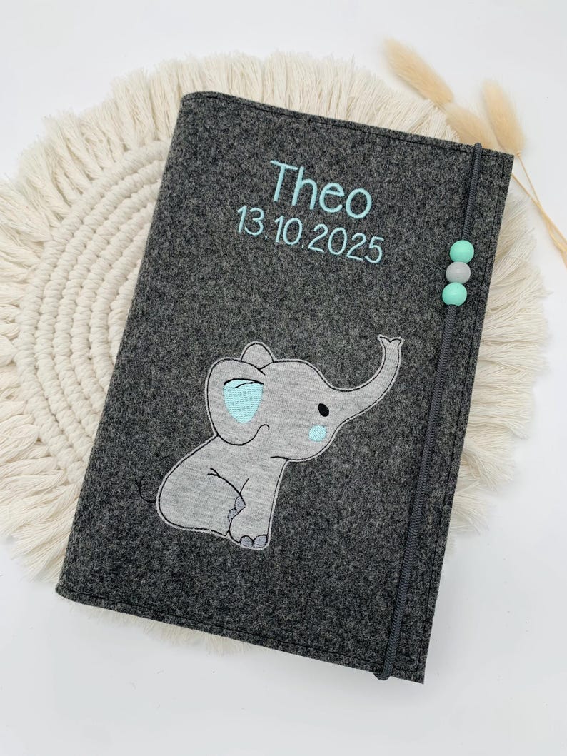 May include: A dark gray felt notebook cover featuring an embroidered light gray elephant with light blue accents. The cover is personalized with the name Theo and the date 13.10.2025 in light blue thread. A gray elastic band with three small beads secures the cover.