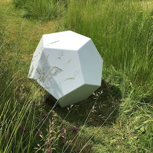 May include: A white, geometric, multi-sided sculpture sits in a field of green grass.