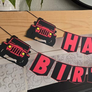 Könnte beinhalten: Ein festliches Geburtstagsbanner mit roten Buchstaben auf schwarzen Wimpeln, die "HAPPY BIRTHDAY" buchstabieren. Das Banner ist mit roten Jeep-Ausschnitten verziert. Das Banner ist über einem hölzernen Kaminsims gespannt.