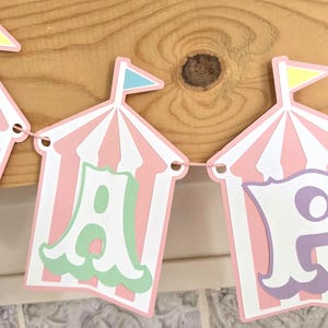May include: A pink and white striped banner with the word "Happy" spelled out in colorful letters. Each letter is inside a pink and white striped tent with a small flag on top.