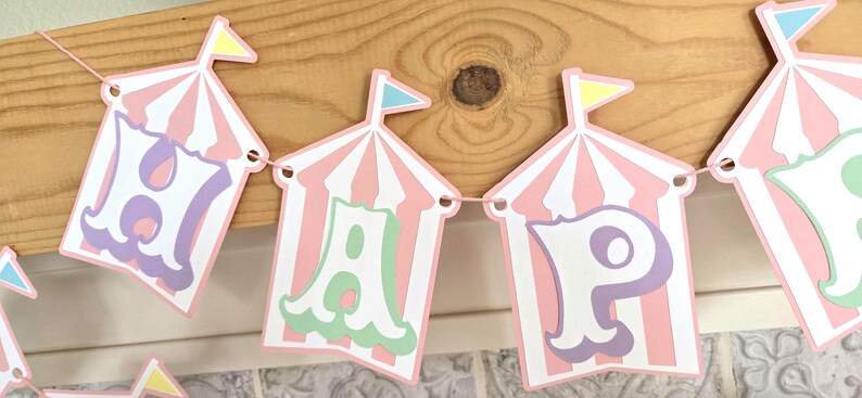 May include: A pink and white striped banner with the word "Happy" spelled out in colorful letters. Each letter is inside a pink and white striped tent with a small flag on top.