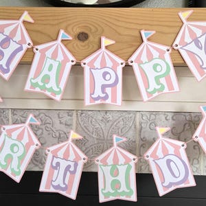 May include: A pink and white circus tent themed banner with the words "Happy Birthday" in pastel colors.