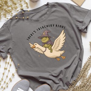 Peut inclure: T-shirt gris avec une illustration fantaisiste d'une grenouille portant un chapeau et une cape de sorcier, chevauchant une oie. Le texte "THERE'S TREACHERY AFOOT" est au-dessus. Le t-shirt est associé à des baskets grises.