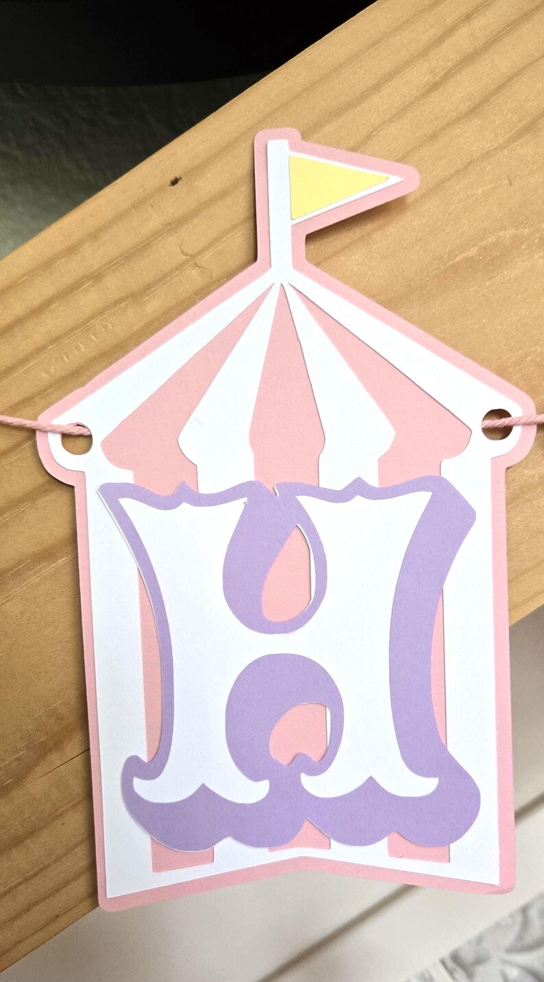 May include: Pink and white circus tent banner with a purple letter H. The banner is strung with white twine.
