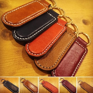 May include: Five leather keychains in different colors, each with a stitched edge and a metal ring. The keychains are arranged in a stack, with the brown keychain on top and the burgundy keychain on the bottom.