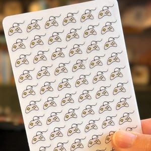 May include: A white card featuring a pattern of small, beige video game controller icons with black outlines. The card has the text "1801 @co." and "Order More:" with a QR code. The card is held in a person's hand.
