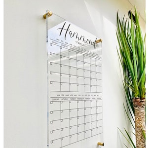 May include: A clear acrylic calendar with a black grid and the text "Hammond Family" at the top. The calendar is mounted on a white wall with gold-colored mounting hardware.