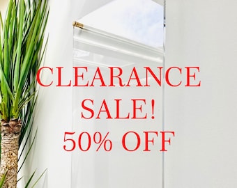 Może przedstawiać: Przezroczysty znak akrylowy z złotymi metalowymi wspornikami wiszący na białej ścianie. Znak zawiera napis "CLEARANCE SALE! 50% OFF" w czerwonym kolorze.
