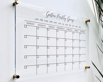 May include: A clear acrylic calendar with gold-colored hardware. The calendar grid is printed in black, with the text "Gallow Reality Group" at the top. Months and days of the week are labeled. The calendar is mounted on a white wall.